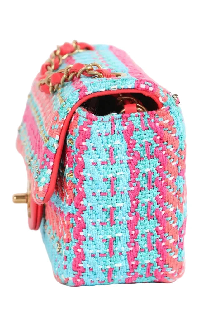 A close-up of a woven handbag in bright pink and light blue tweed with gold-tone chain detailing and a gold clasp, shown on a plain white background.