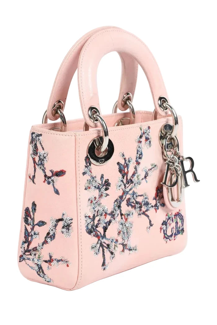 A light pink leather Lady Dior handbag with floral embroidery, silver-tone hardware, and a hanging 'Dior' charm, shown on a white background.