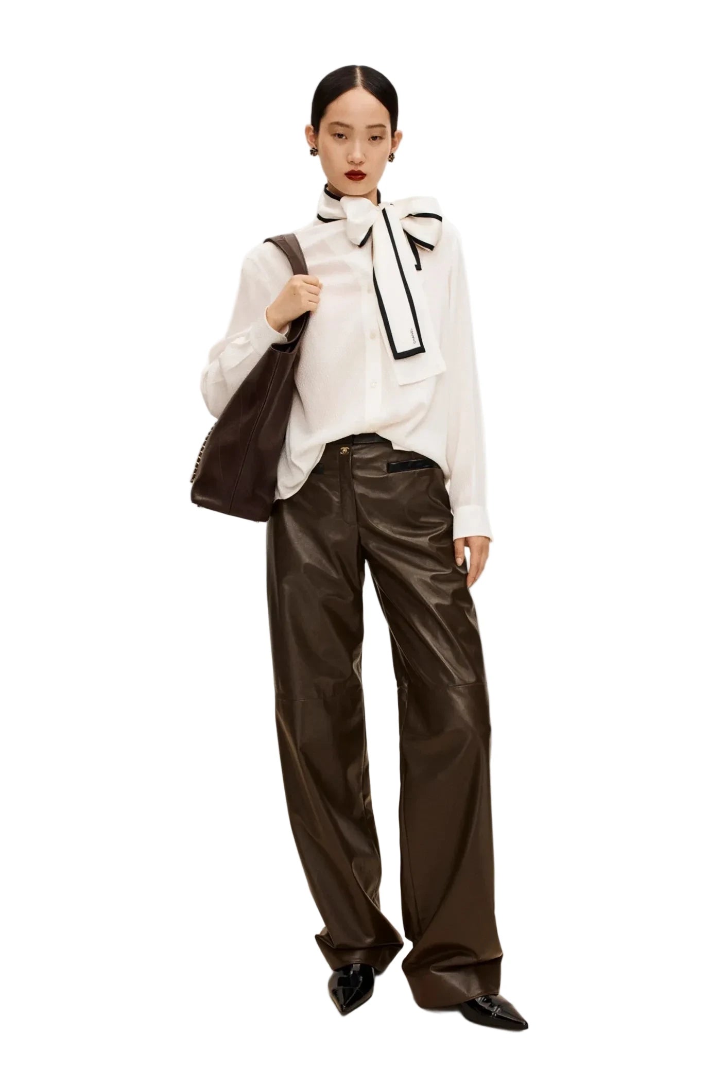 Woman wearing a white blouse with a black bow and dark pants, holding a black bag on a beige background