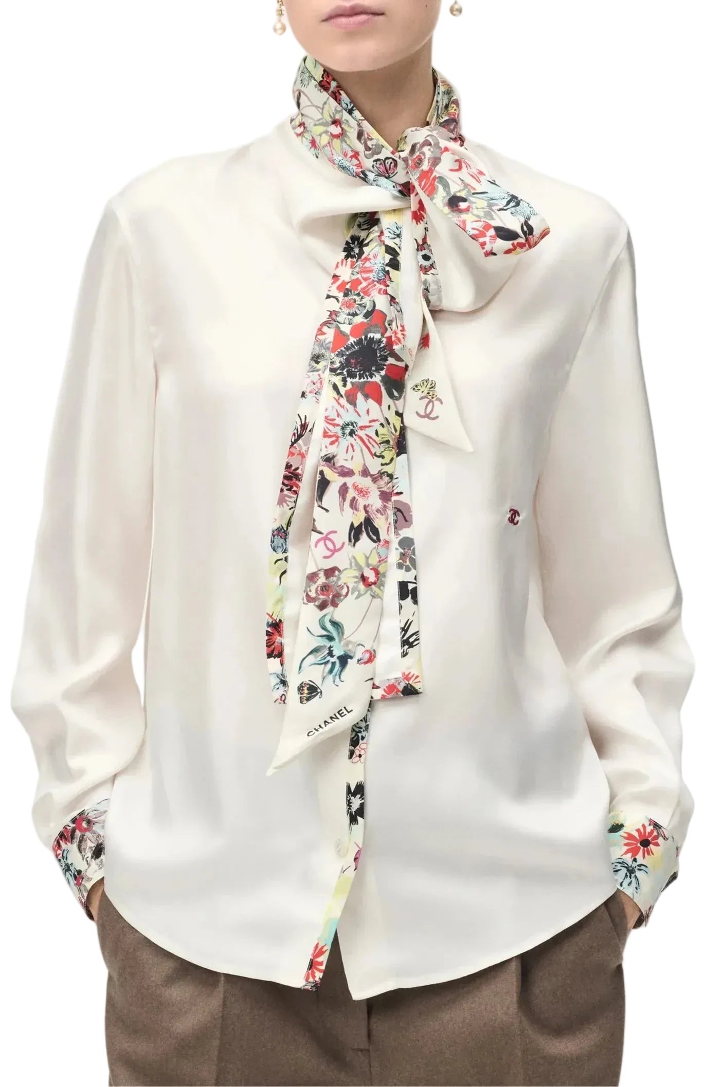 A woman wearing a white long-sleeve blouse with a colorful floral silk neck scarf tied around the collar, and matching floral cufflinks on the sleeves. The blouse has a small red embroidered Chanel logo on the left chest.