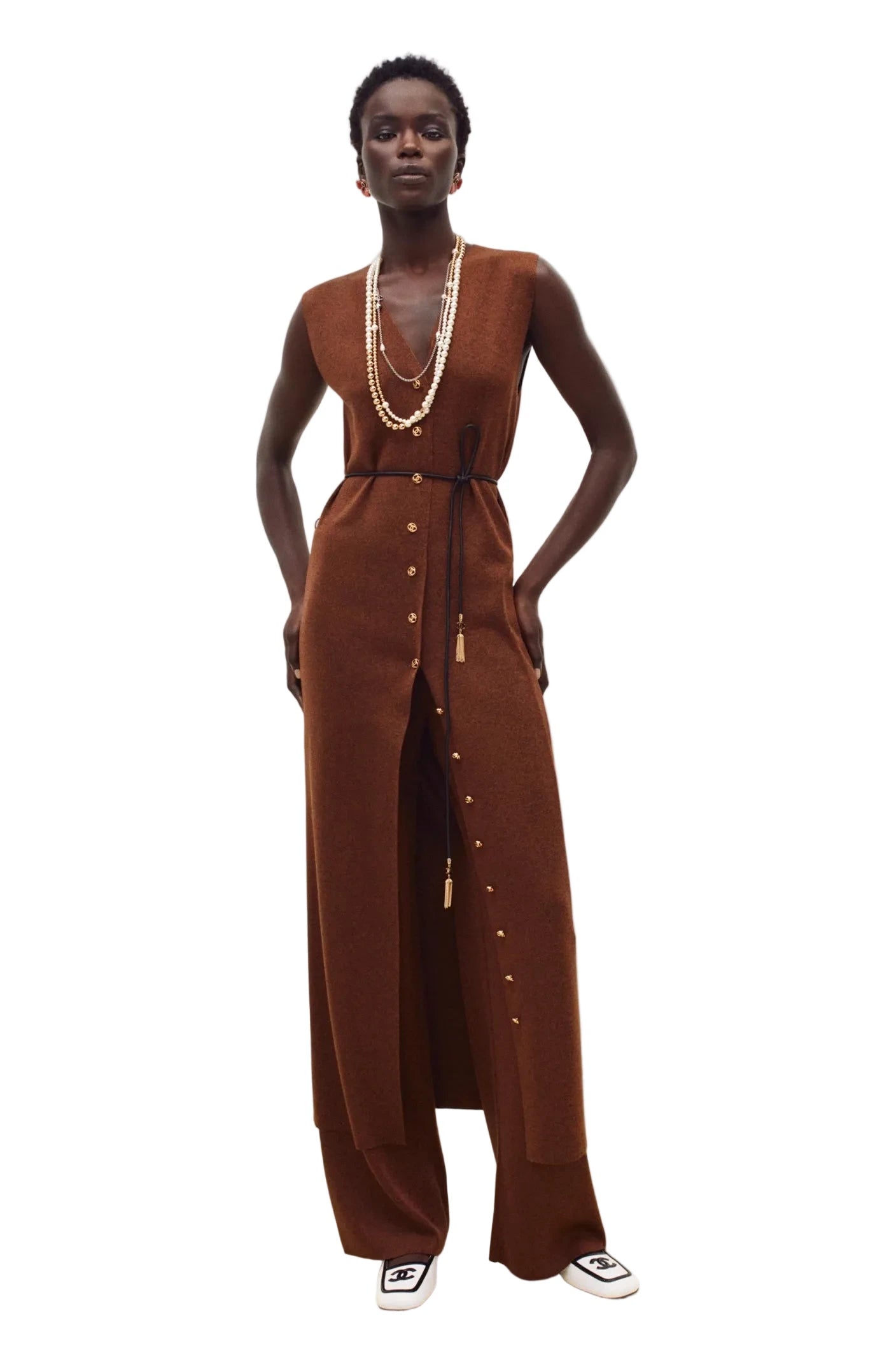 A model stands against a plain off-white studio background wearing a sleeveless, wide-leg brown jumpsuit with a front button closure and tassel tie detail. She is also wearing layered gold necklaces and white slip-on shoes.
