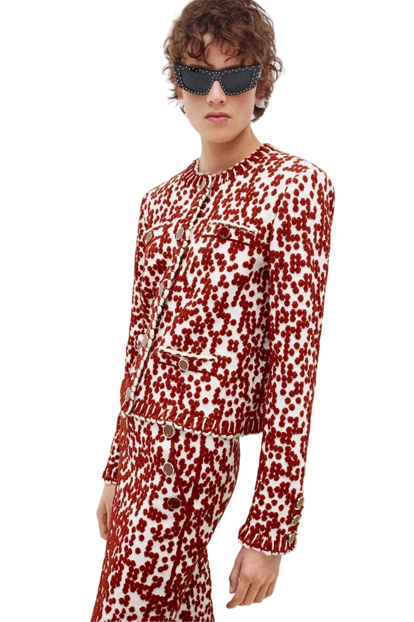A model wearing a matching red and white patterned tweed jacket and trousers set, styled with dark sunglasses, against a plain white studio background.