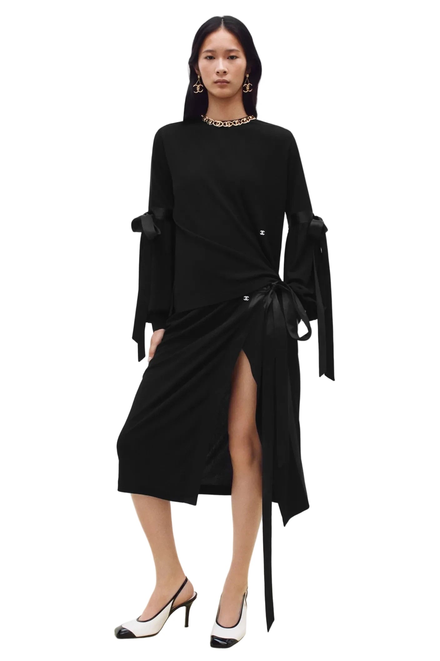 A model wearing a black long-sleeve wrap-style midi dress with a high thigh slit, tie waist, and draped sleeves, paired with black and white heels against a plain white studio background.