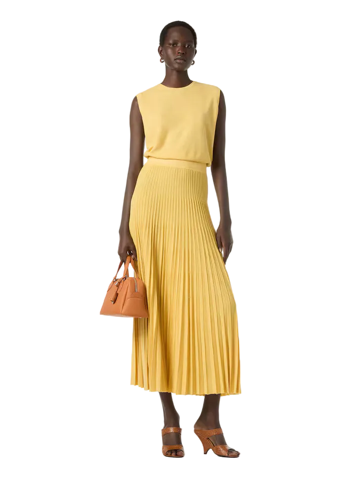 Woman wearing a yellow sleeveless top and pleated skirt holding a brown handbag on a white background