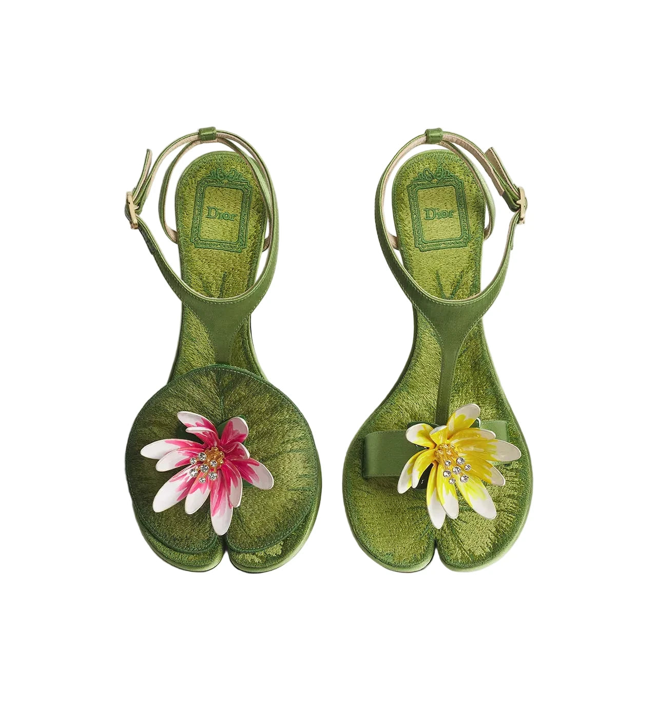 A pair of green tabi-style sandals with floral embellishments, displayed on a light gray background. The sandals feature olive green leather with a textured pattern, a gold-tone ankle strap, and decorative flowers on the toe area (one pink and white, one yellow and white).