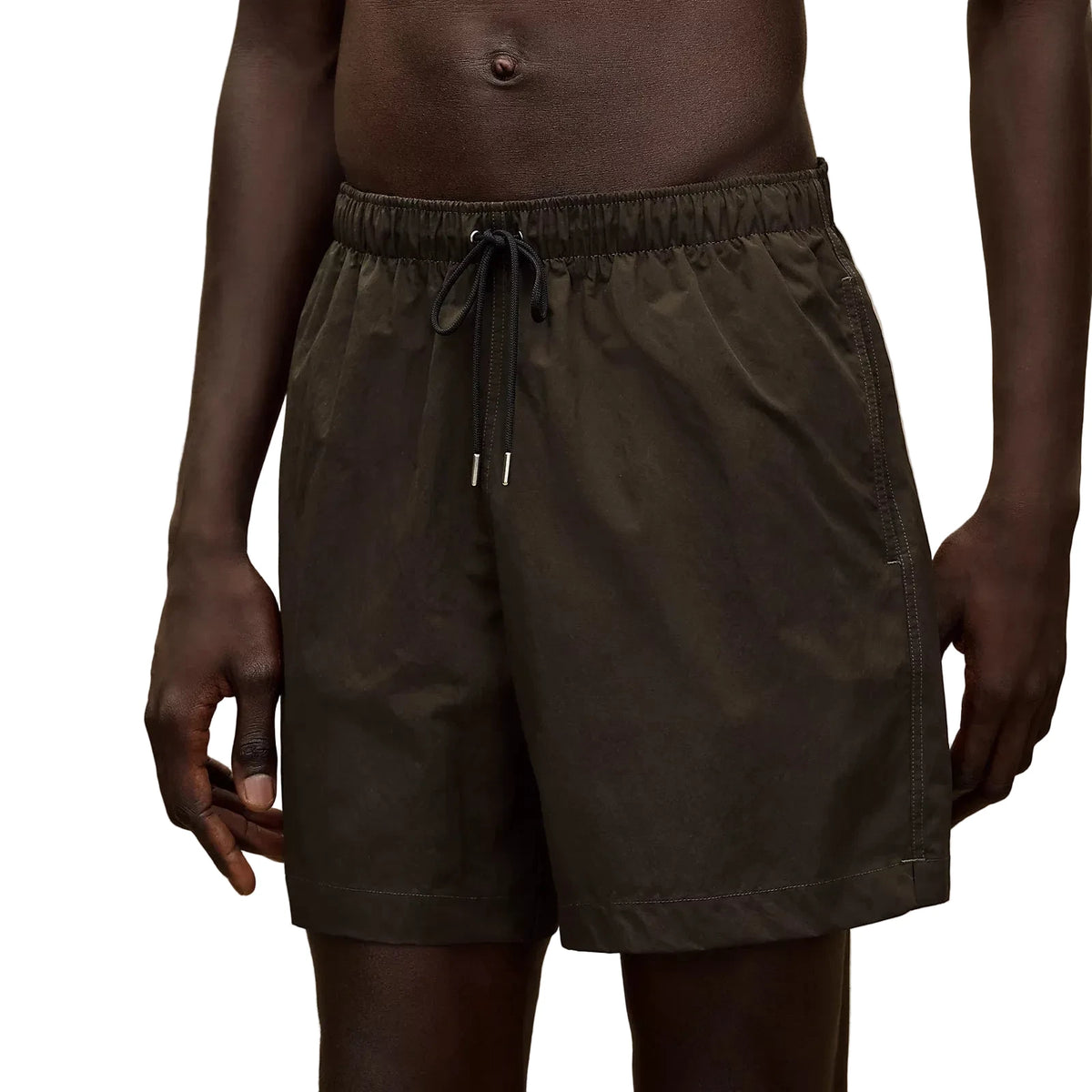A close-up of a person wearing dark olive green drawstring shorts against a plain beige studio background.