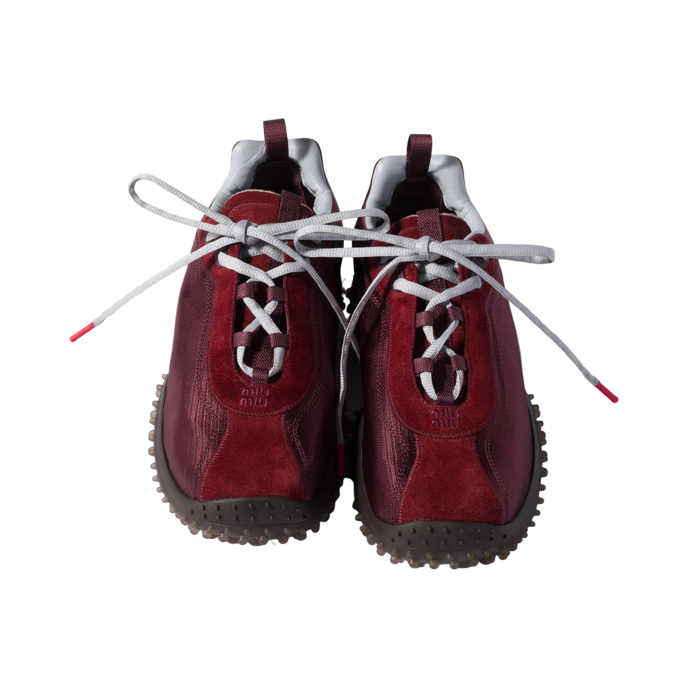 A pair of low-top sneakers in deep burgundy suede and leather, featuring white laces knotted in the center, a white collar lining, and dark gray spiked outsoles, photographed from a top-down angle on a light beige background.