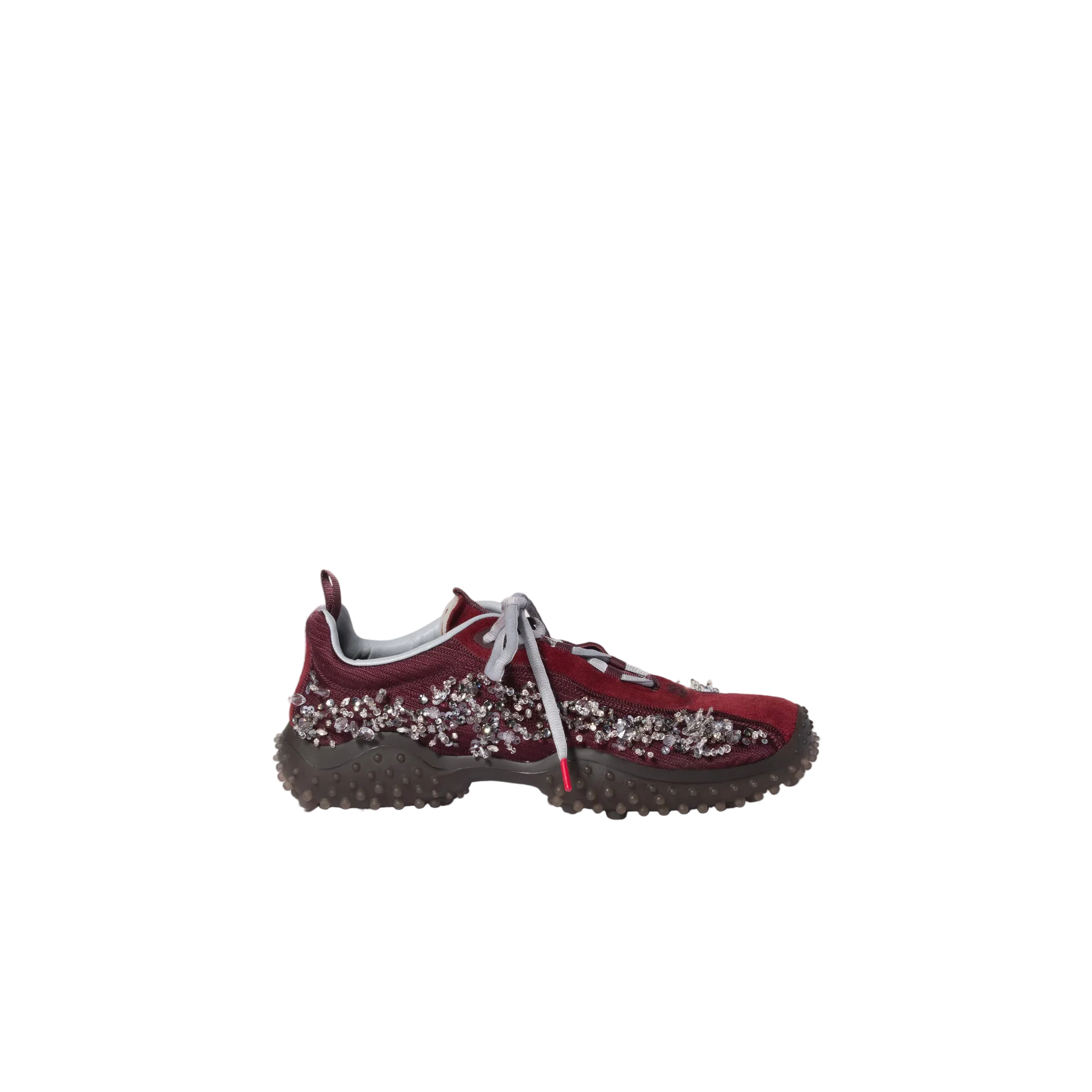 A single low-top sneaker shown in profile against a solid light beige background. The shoe features a deep burgundy suede upper embellished with silver rhinestones, a chunky dark brown textured sole, and light gray laces.