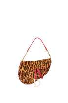 A Dior saddle bag in leopard print with a red leather strap and a gold-tone Dior logo charm, photographed on a clean white background.