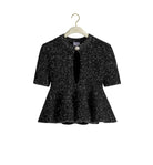A black short-sleeve peplum jacket with a white speckled pattern, hanging on a light wood hanger with a gold hook against a plain white background.