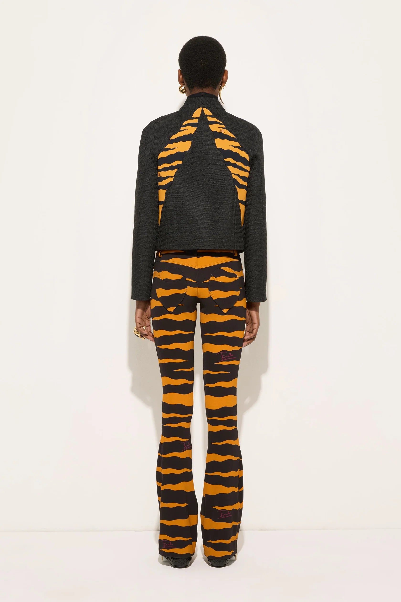A model shown from the back wearing a black cropped jacket with a bold orange and black tiger-stripe pattern on the back, paired with matching patterned flared pants, against a plain white studio background.