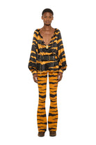 A model stands against a plain white studio background wearing a matching tiger-print two-piece set consisting of a long-sleeve top with a black belt and flared pants.