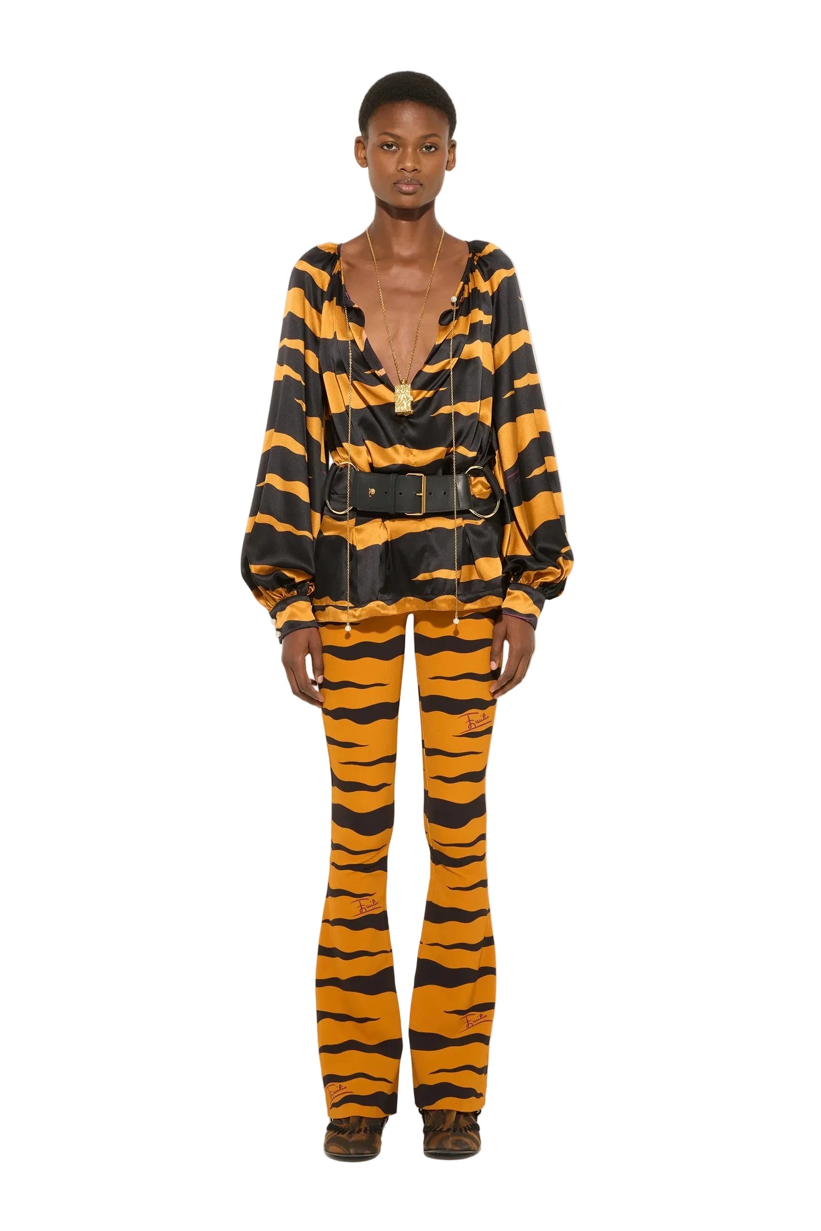 A model stands against a plain white studio background wearing a matching tiger-print two-piece set consisting of a long-sleeve top with a black belt and flared pants.
