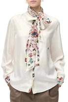 A woman wearing a white long-sleeve blouse with a colorful floral silk neck scarf tied around the collar, and matching floral cufflinks on the sleeves. The blouse has a small red embroidered Chanel logo on the left chest.