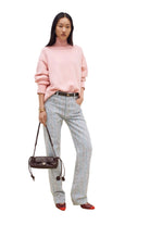 A model stands against a plain off-white studio background wearing a light pink turtleneck sweater, light blue and white vertically striped trousers, a thin black belt, and red pointed flats. She is holding a dark brown quilted shoulder bag.