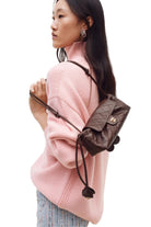 A woman wearing a pink knit sweater and patterned pants carries a small dark brown quilted shoulder bag with a gold-tone logo clasp and drawstring detail against a plain light background.