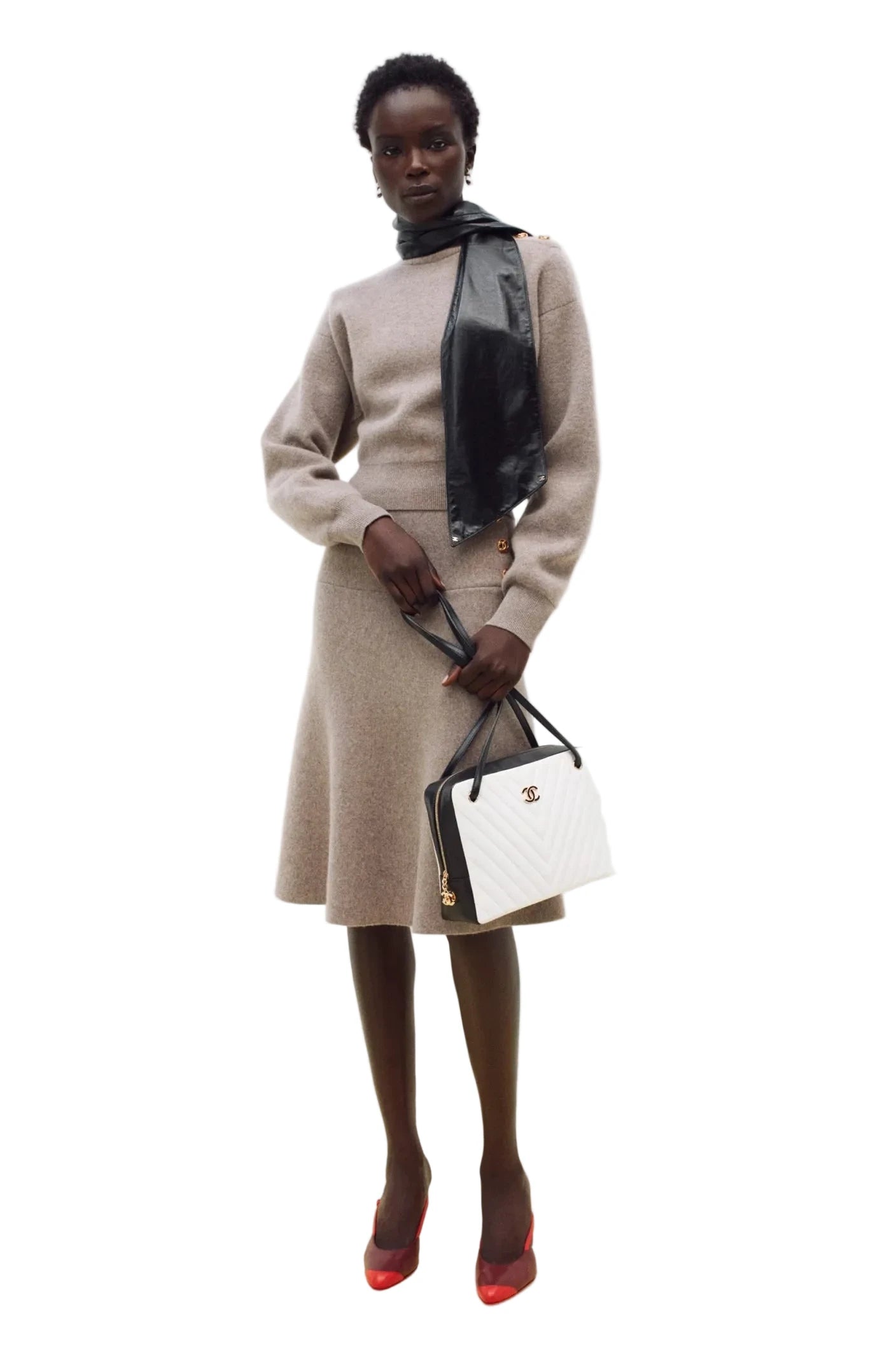 A model stands against a plain white studio background wearing a beige knit turtleneck sweater and matching knee-length skirt, with a black scarf and a white quilted handbag with black trim.
