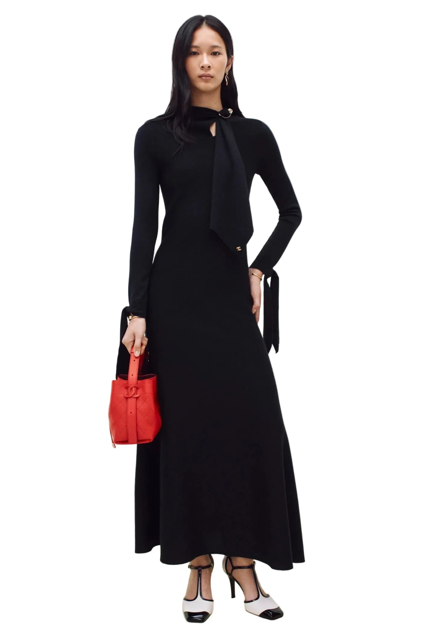 A model wearing a long-sleeved black knit maxi dress with a high neck and a large bow detail at the collar, holding a small red handbag. She is wearing black and white heels against a clean white studio background.