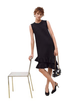 A woman models a sleeveless black midi dress with a ruffled hem, standing next to a minimalist metal chair and holding a small black floral handbag.