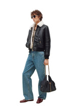 A model wearing a black leather aviator jacket with a white shearling collar, blue wide-leg jeans, burgundy loafers, and holding a black quilted handbag with a gold chain strap, set against a plain white studio background.