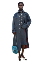 A model stands against a plain white studio wall wearing a long blue denim button-front coat over a dark dress, holding a small blue quilted shoulder bag.