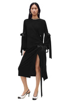 A model wearing a black long-sleeve wrap-style midi dress with a high thigh slit, tie waist, and draped sleeves, paired with black and white heels against a plain white studio background.