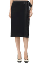 A model wearing a black wrap midi skirt with a side tie and a small silver logo charm, paired with black and white pointed-toe heels, against a plain white studio background.