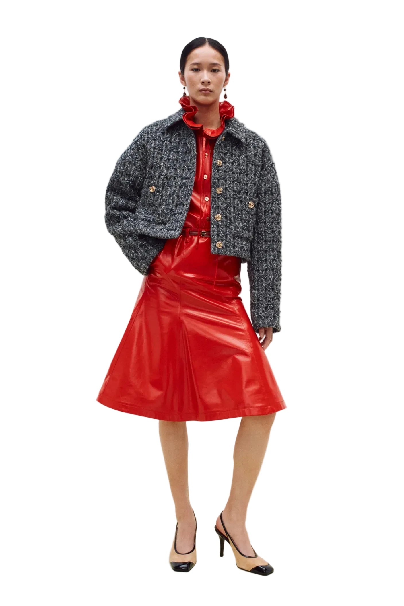 A model stands against a plain white studio wall wearing a gray and black tweed cropped jacket over a bright red ruffled blouse and a shiny red skirt, with beige-and-black heels.