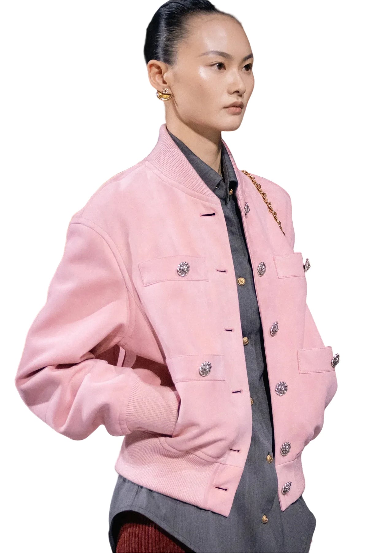 A model on a runway wearing a light pink structured jacket with decorative silver-tone embellishments, layered over a dark gray shirt.