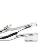 A close-up side view of a metallic silver slingback shoe with a low block heel and a small gold logo detail on the side, set against a plain white background.