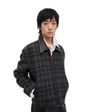 A model wearing a dark gray and black plaid zip-up jacket over a white collared shirt, photographed against a clean white studio background.