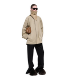 A model stands against a white studio background wearing an oversized beige jacket with a high mock neck, black wide-leg trousers, dark brown fuzzy boots, and a brown textured shoulder bag.
