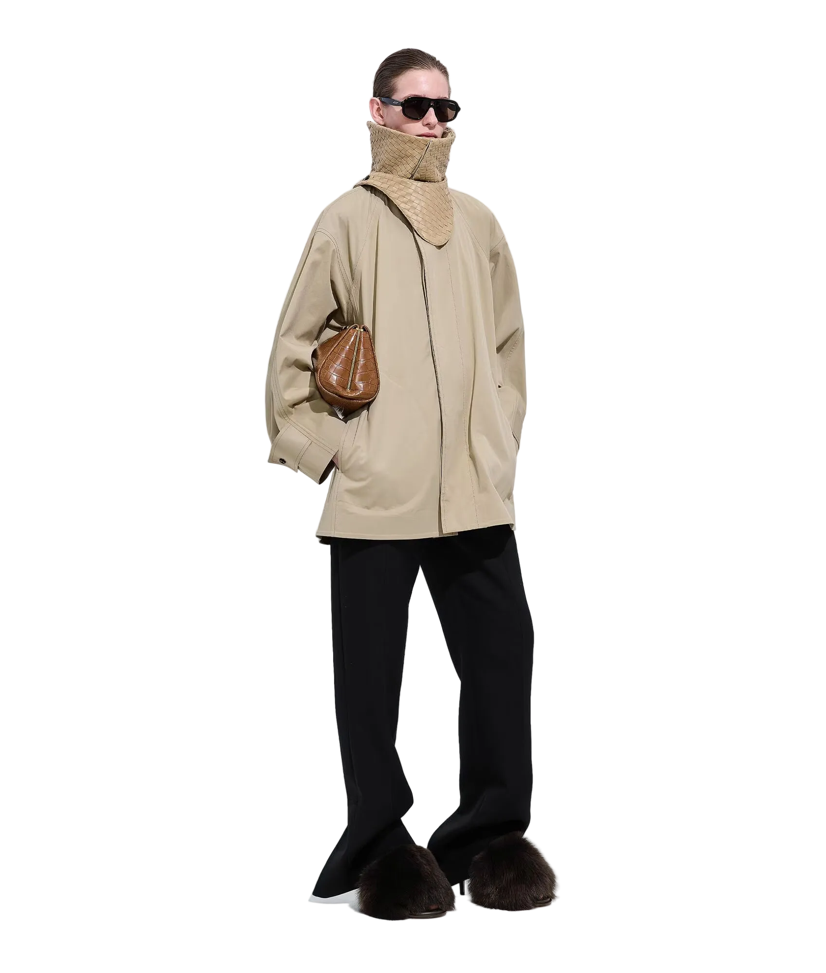 A model stands against a white studio background wearing an oversized beige jacket with a high mock neck, black wide-leg trousers, dark brown fuzzy boots, and a brown textured shoulder bag.