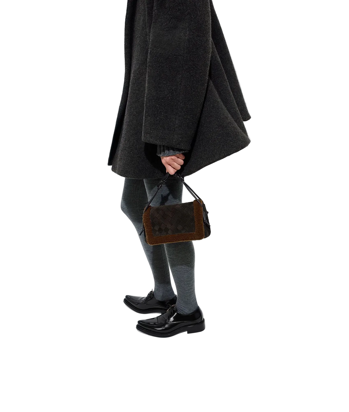 A person wearing a dark charcoal coat, gray tights, and black leather loafers holds a brown textured handbag with a dark strap against a clean white studio background.