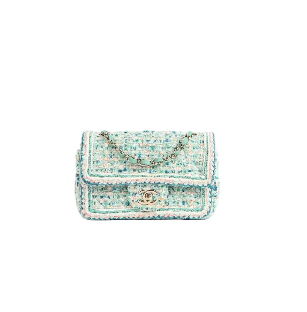 A small tweed shoulder bag in light blue, white, and beige with a gold-tone chain strap and a gold interlocking CC logo on the front flap, photographed on a clean white background.