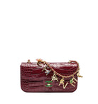 A burgundy crocodile-embossed leather handbag with gold-tone hardware and a decorative charm strap featuring interlocking 'C' and 'V' logos, a bell, and colorful enamel details, set against a plain white background.