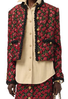 A cropped, long-sleeve jacket with a red and green floral pattern, black lace trim, and gold buttons, styled over a cream-colored button-down shirt.