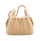 A beige quilted suede drawstring handbag with fluffy cream shearling lining, gold-tone chain handles, and a gold interlocking CC logo on the front, photographed against a plain white background.