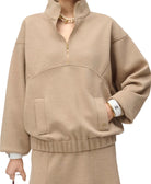 A close-up of a person wearing a beige quarter-zip pullover sweatshirt with a stand collar, front patch pockets, and a matching ribbed hem, paired with a matching skirt. The model is also wearing a gold necklace, a white cuff bracelet, and a gold watch.