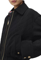 Close-up of a black quilted jacket with gold-tone zipper and gold double-C logo buttons, worn by a model. The jacket features a pointed collar, a diamond-quilted pattern, and a pocket on the lower left side.