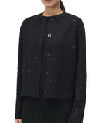 A close-up of a model wearing a black cropped cardigan jacket with textured bead embellishments and silver-tone buttons, paired with a matching black skirt against a plain light background.
