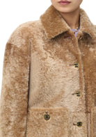 A close-up of a person wearing a plush faux-fur or shearling-style jacket in warm tan and beige tones, with dark buttons featuring a small interlocking CC logo. The collar and inner lining are a slightly darker tan, and the visible inner layer at the neckline appears light purple.