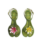 A pair of green tabi-style sandals with floral embellishments, displayed on a light gray background. The sandals feature olive green leather with a textured pattern, a gold-tone ankle strap, and decorative flowers on the toe area (one pink and white, one yellow and white).