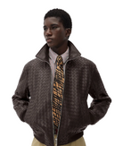 A male model wearing a dark brown woven leather bomber jacket over a striped shirt and a patterned tie, photographed against a light gray studio background.