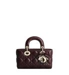 A dark burgundy quilted leather handbag with gold-tone hardware, featuring a top handle, circular ring detail, and a hanging Dior logo charm, set against a plain light gray background.
