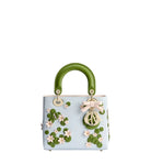 A light blue leather handbag with a glossy green top handle, decorated with 3D green lily pads, pink flowers, and a pink bow with a gold-tone Christian Dior logo charm.