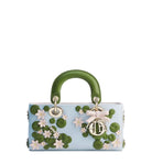 A light blue leather handbag with a structured top handle, decorated with 3D green lily-of-the-valley motifs, white flowers, and a gold-tone Christian Dior charm with a pink bow, set against a plain light gray background.