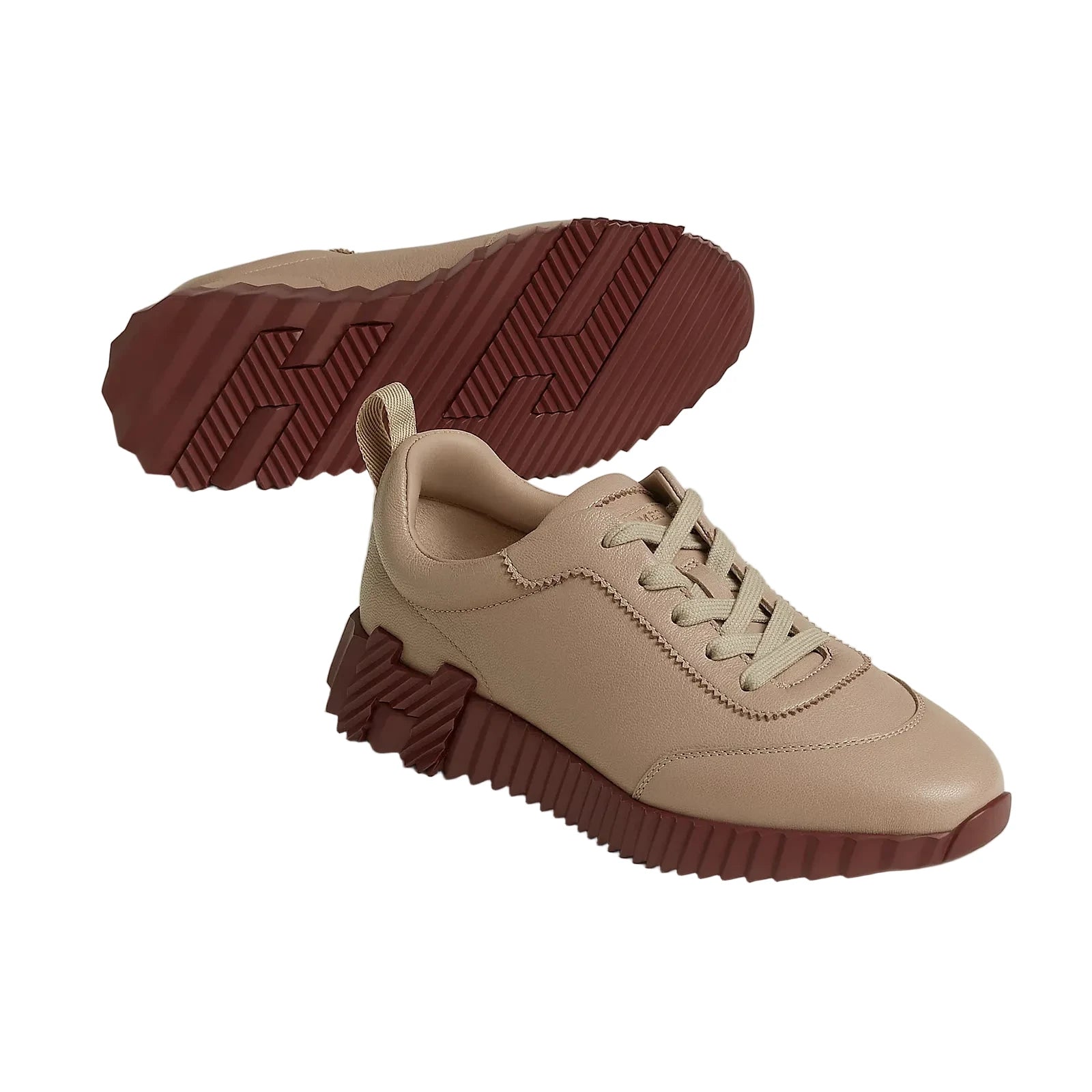 A pair of low-top sneakers in beige with a contrasting dark reddish-brown sole and outsole, shown on a light beige studio background. One shoe is positioned upright, and the other is shown from the side profile with its sole facing up to display the textured tread pattern.