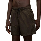 A close-up of a person wearing dark olive green drawstring shorts against a plain beige studio background.