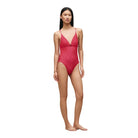 A model stands barefoot against a warm beige studio backdrop wearing a one-piece swimsuit with a red and pink floral pattern.